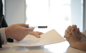 Two pairs of hands: one holding a pen and papers, and the other waiting to sign.